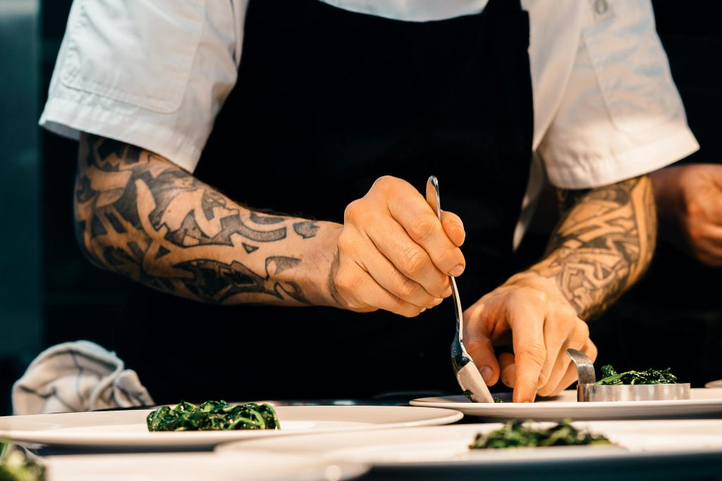 Chef privado en Marbella A tattooed chef carefully plates a spinach dish in a professional kitchen setting, showcasing culinary expertise.