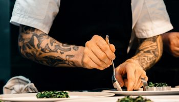 A tattooed chef carefully plates a spinach dish in a professional kitchen setting, showcasing culinary expertise.